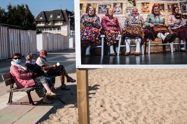 Люди в защитных на пляже возле фотографии из серии «Во имя матери» фотографа Нади Феррухи, представленной в Ульгате, на северо-западе Франции - Sputnik Кыргызстан