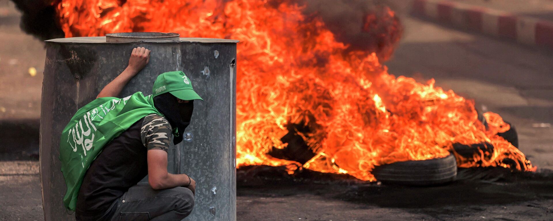 Палестинский протестующий в маске укрывается возле горящих шин во время столкновений с израильскими силами в городе Хеврон - Sputnik Кыргызстан, 1920, 16.10.2023
