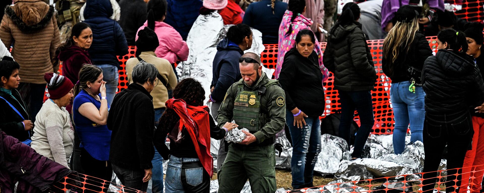 Офицер таможни и пограничной службы США (в центре) раздает еду иммигрантам, ожидающим оформления в транзитном центре пограничного патруля США, после пересечения границы с Мексикой в ​​Игл-Пасс, штат Техас - Sputnik Кыргызстан, 1920, 27.01.2024