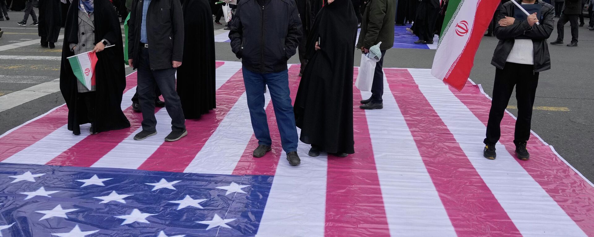 Иранцы стоят на баннере с изображением флага США - Sputnik Кыргызстан, 1920, 18.03.2026